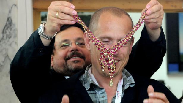 Ein Mann hält eine Halskette mit roten Steinen vor den Hals eines anderen Mannes.