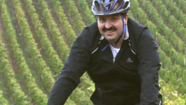 Ein Mann mit Schnurrbart fährt mit dem Fahrrad durch einen Weinberg.