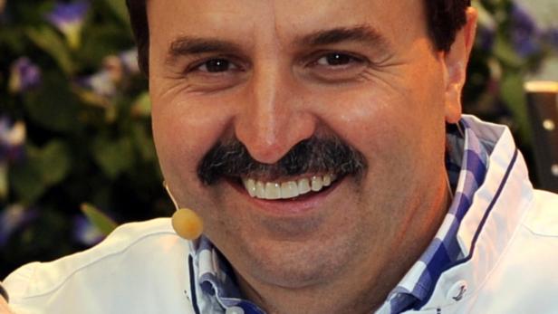 Ein lächelnder Mann mit Schnurrbart und Mikrofon am Kragen.