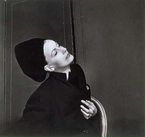 Greta Garbo in einem eleganten schwarzen Outfit und Hut, den Kopf nach oben geneigt.