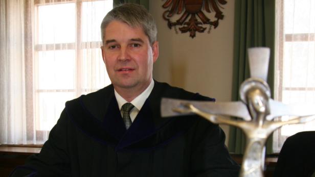 Ein Mann in einer Robe sitzt vor einem Fenster und einem Kruzifix.