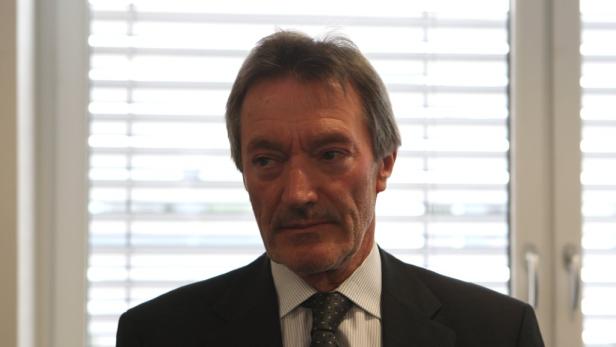 Ein Mann in Anzug und Krawatte vor einem Fenster mit Jalousien.