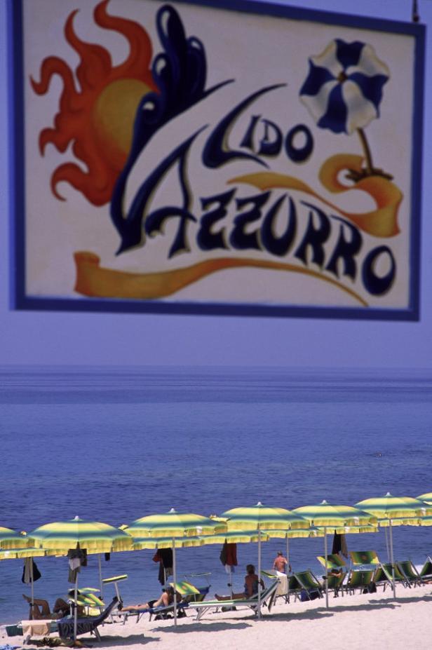 Ein Strand mit gelb-grünen Sonnenschirmen und dem Schild „Lido Azzurro“ im Hintergrund.