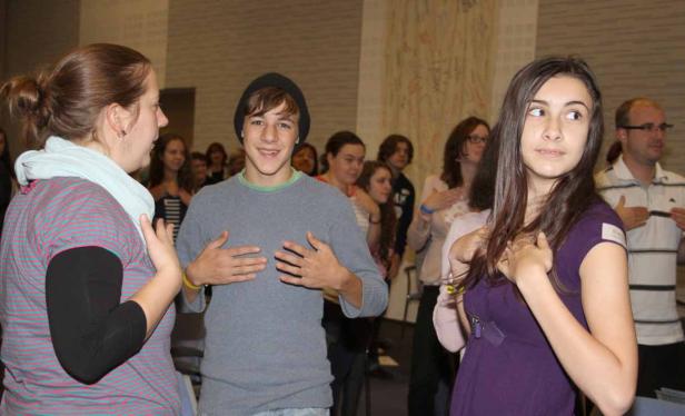 Eine Gruppe von Menschen steht in einem Raum und hält die Hände auf die Brust.