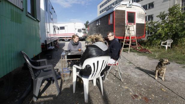 Drei Personen sitzen an einem Tisch vor Bauwagen, während ein Hund zusieht.