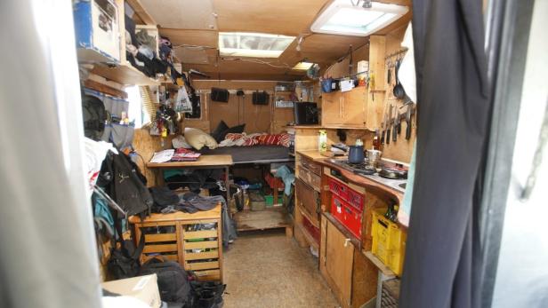 Blick in das hölzerne Innere eines kleinen Wohnmobils mit Bett, Küche und Stauraum.