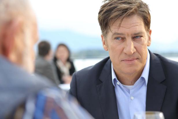 Ein Mann in einem blauen Anzug sitzt an einem Tisch und spricht mit anderen Personen.