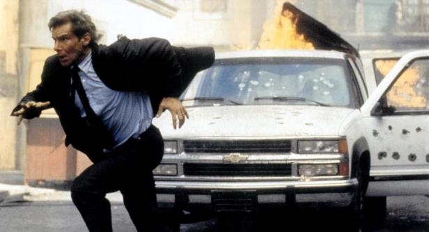 Harrison Ford rennt vor einem brennenden und von Kugeln durchlöcherten Auto weg.