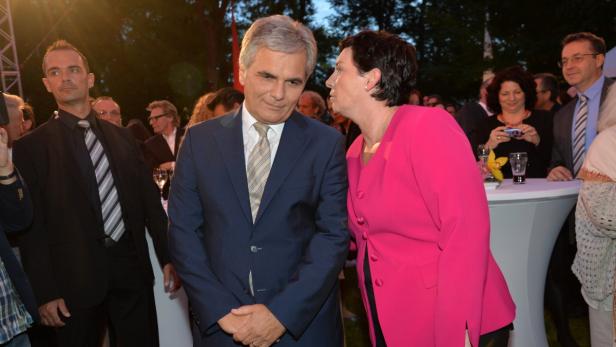 Eine Frau im pinkfarbenen Blazer flüstert einem Mann im Anzug etwas ins Ohr.