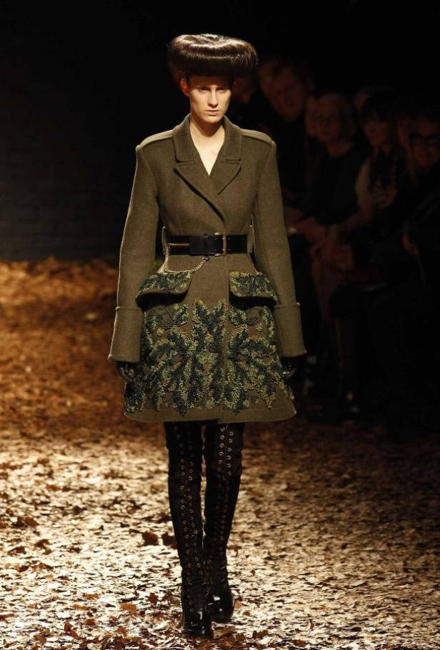 Eine Frau in einem grünen Mantel mit Blattmuster und hohen Stiefeln auf einem Laufsteg.