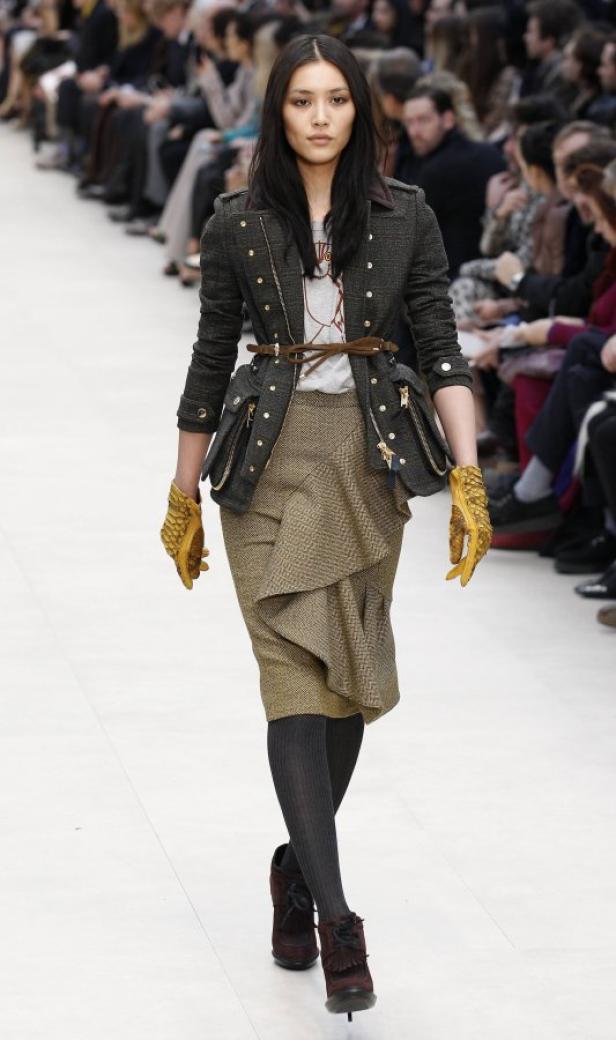 Eine Frau präsentiert auf einem Laufsteg eine Jacke mit goldenen Handschuhen und einen Rock.