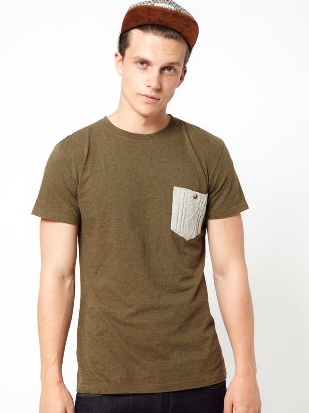 Ein junger Mann mit brauner Mütze und olivgrünem T-Shirt mit Stricktasche posiert vor einem weißen Hintergrund.