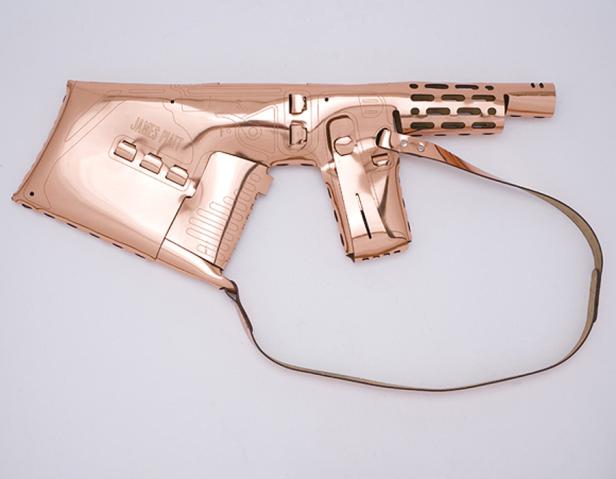 Eine roségoldene, flache Gewehrskulptur mit dem Namen „James Platt“ liegt auf einem weißen Hintergrund.