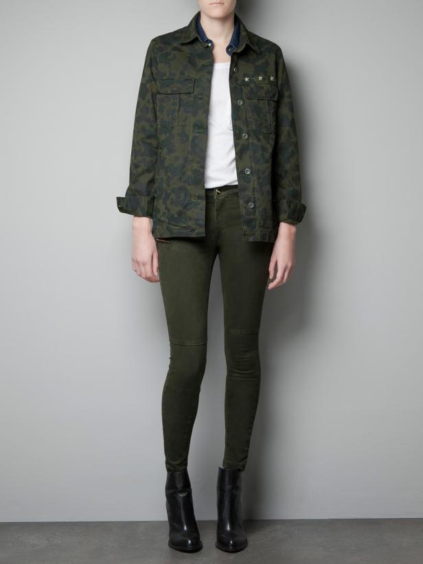 Eine Frau trägt eine grüne Camouflage-Jacke, eine weiße Bluse, grüne Jeans und schwarze Stiefeletten.
