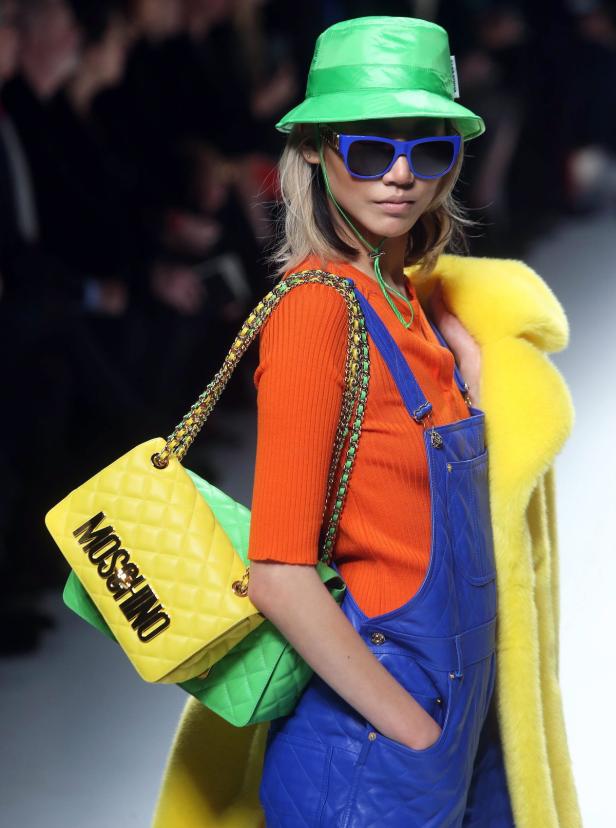 Ein Model mit Hut, Sonnenbrille und farbenfroher Kleidung präsentiert eine Moschino-Tasche.