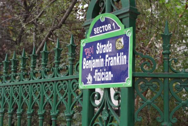 Ein Straßenschild „Strada Benjamin Franklin“ im Sektor 1, befestigt an einem grünen Metallzaun.
