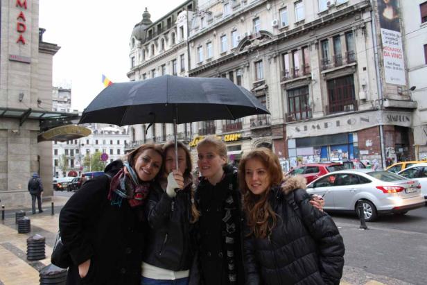 Vier Frauen stehen unter einem Regenschirm in einer städtischen Umgebung.