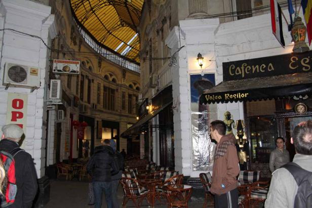 Menschen besuchen ein Café in einer belebten Gasse mit einem Glasdach.