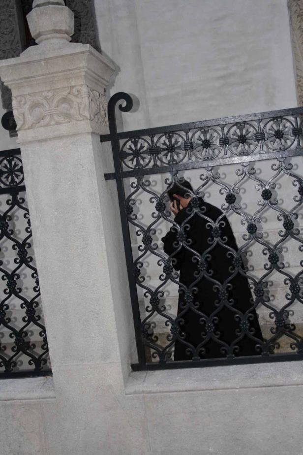 Eine Person in dunkler Kleidung telefoniert hinter einem verzierten Eisenzaun.