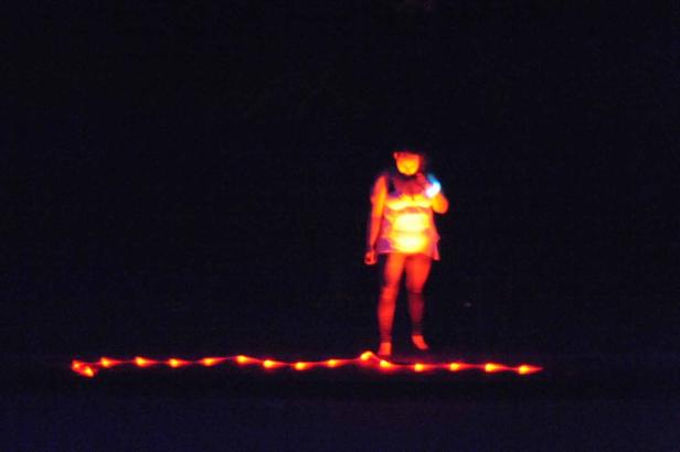 Eine Frau steht im Dunkeln, beleuchtet von Lichtern an ihrer Kleidung und einer Lichterkette am Boden.