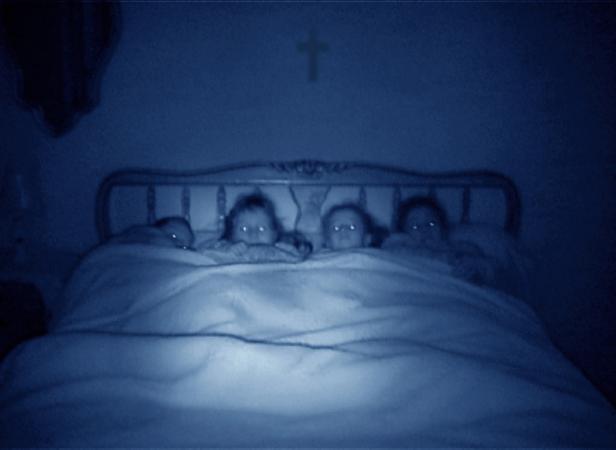 Vier Kinder liegen in einem Bett unter einem Kreuz an der Wand.
