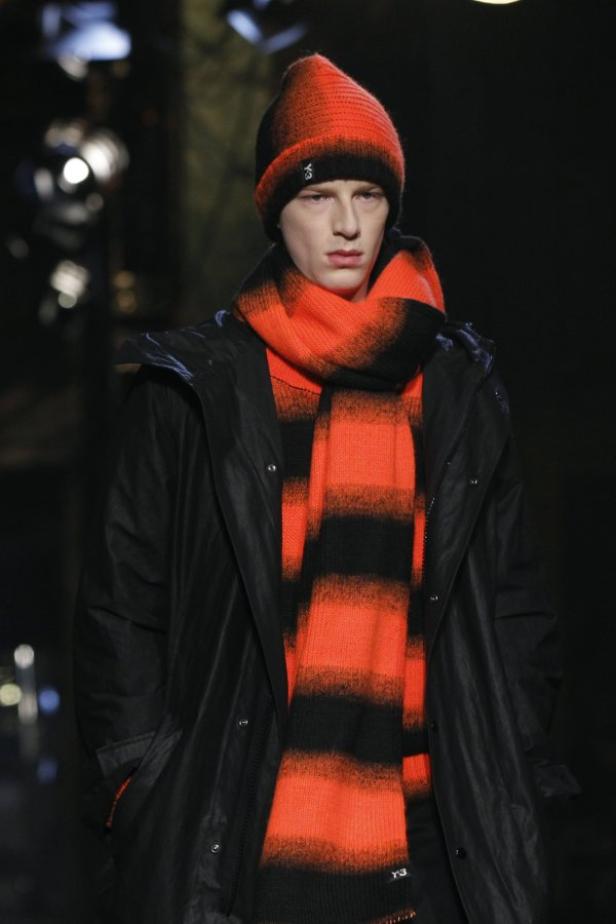 Ein Mann mit Mütze und Schal in Schwarz und Orange trägt einen schwarzen Regenmantel.