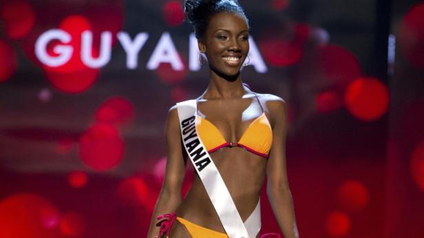Eine lächelnde Frau mit einer Schärpe mit der Aufschrift „Guyana“ vor einem roten Hintergrund.