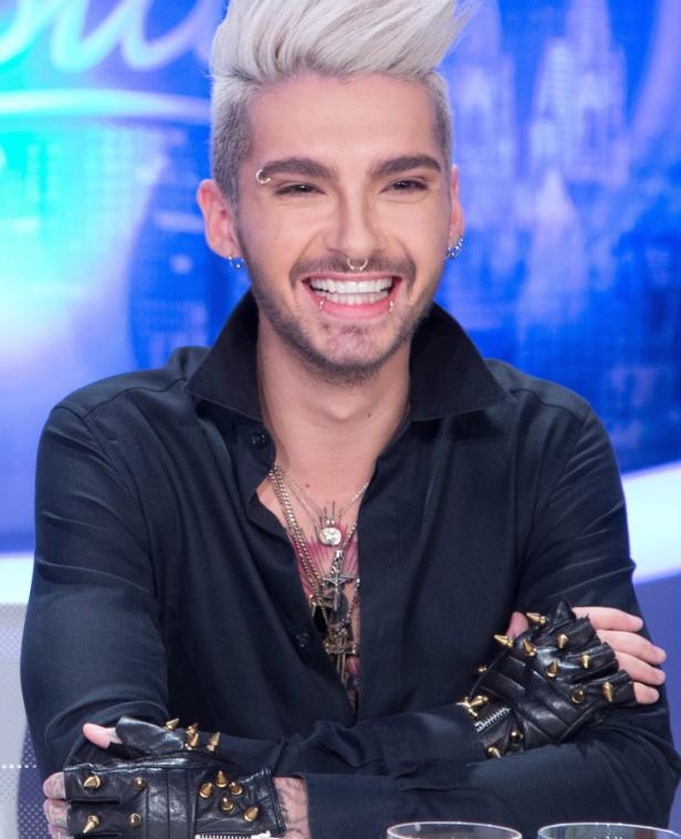 Bill Kaulitz lächelt mit Piercings und Nietenhandschuhen in die Kamera.