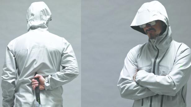 Ein Mann mit Sonnenbrille und Kapuzenjacke, der einen Gegenstand hinter seinem Rücken versteckt.