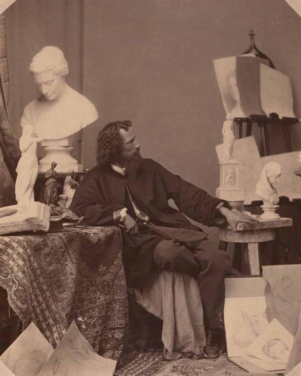 Ein bärtiger Mann sitzt in einem Atelier, umgeben von Skulpturen und Zeichnungen.