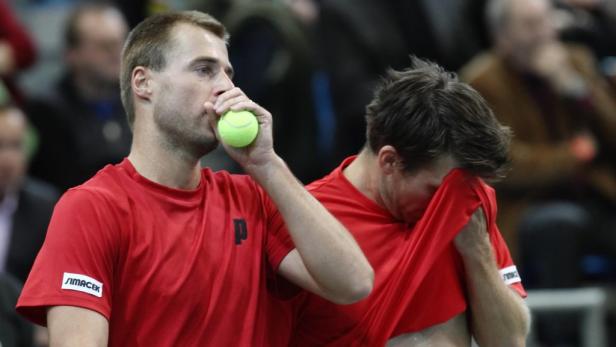 Zwei Tennisspieler in roten Trikots, einer mit einem Tennisball am Mund, der andere verdeckt sein Gesicht mit seinem Trikot.