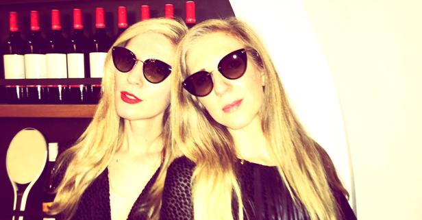 Zwei blonde Frauen posieren mit Sonnenbrillen vor einem Regal mit Weinflaschen.