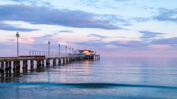 Ein langer Pier mit einem Gebäude am Ende erstreckt sich ins ruhige Meer bei Dämmerung.