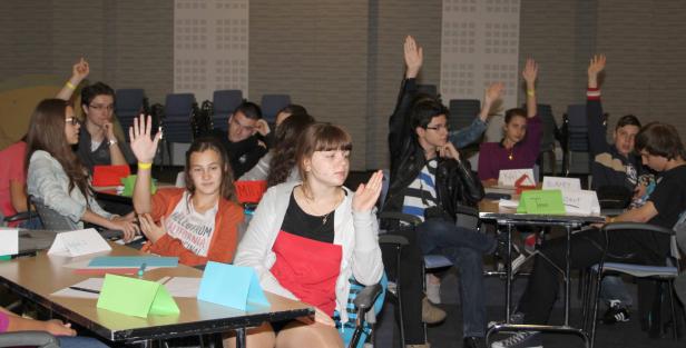 Eine Gruppe von Schülern sitzt an Tischen und hebt die Hände.
