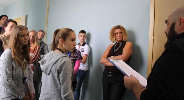 Eine Gruppe von Menschen steht in einem Flur und wartet auf Anweisungen von einer Frau mit einem Klemmbrett.