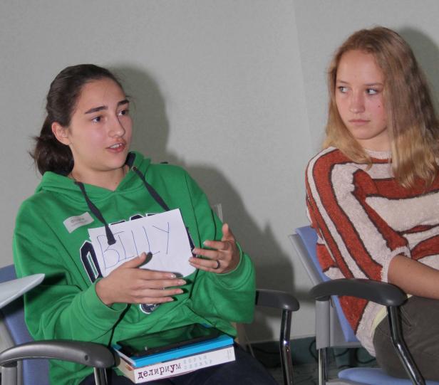 Zwei junge Frauen sitzen auf Stühlen, eine hält ein Schild mit der Aufschrift „Billy“.