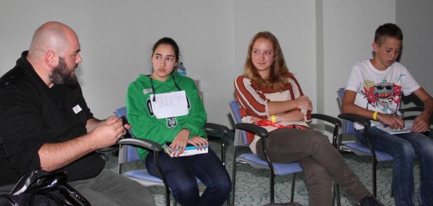 Eine Gruppe von Jugendlichen und ein Mann sitzen auf Stühlen in einem Raum.