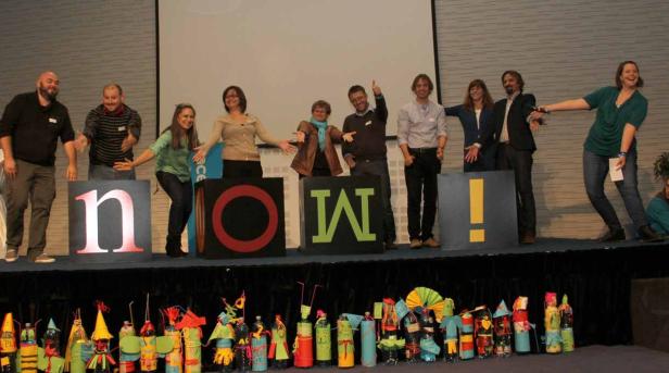 Eine Gruppe von Personen steht auf einer Bühne hinter großen Buchstaben, die das Wort „NOW!“ bilden.