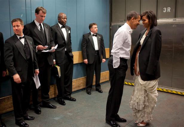 Barack und Michelle Obama stehen sich gegenüber, umgeben von Männern in Anzügen.