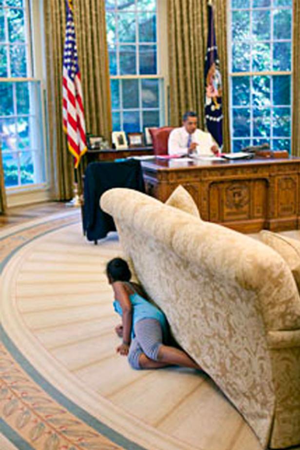 Ein Mädchen versteckt sich hinter einem Sofa im Oval Office, während Barack Obama am Schreibtisch sitzt.