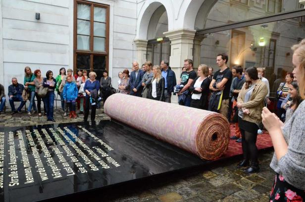 Eine Gruppe von Menschen betrachtet eine Installation mit einer großen Stoffrolle und Text auf einer schwarzen Oberfläche.