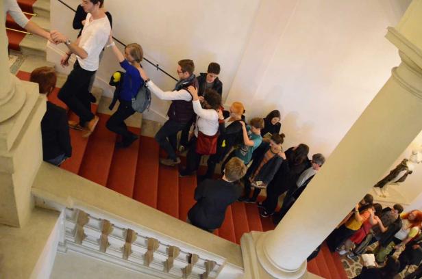 Eine Menschenschlange steigt eine rote Treppe in einem Gebäude hinauf, sich gegenseitig helfend.