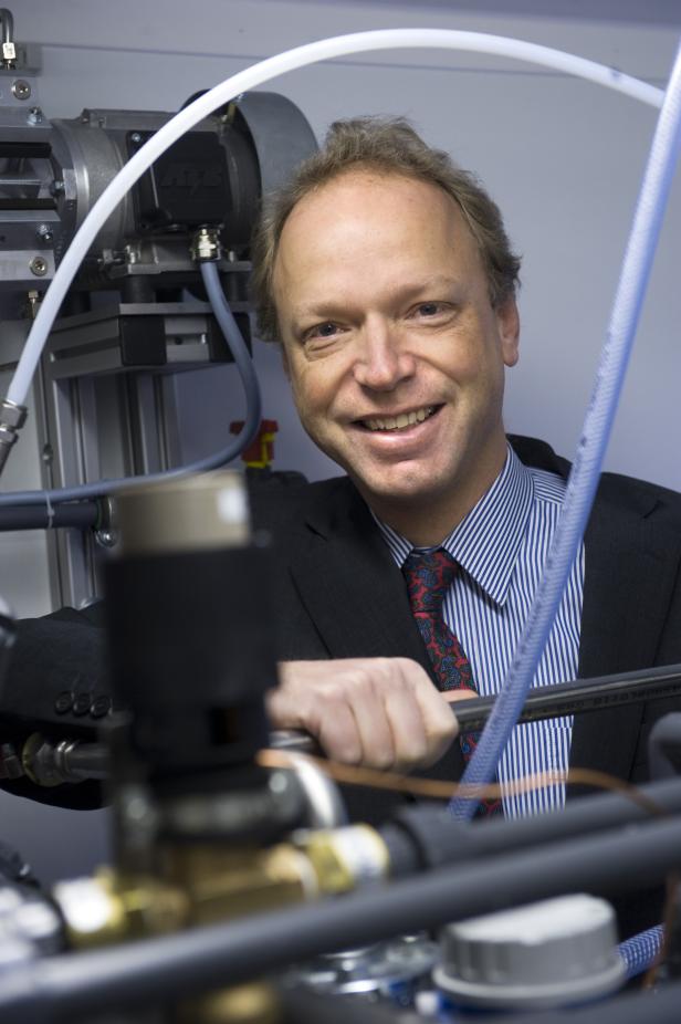 Ein lächelnder Mann im Anzug steht vor einer komplexen technischen Apparatur.