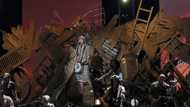 Taylor Swift spielt Banjo auf der Bühne, umgeben von ihrer Band und rustikalen Bühnenelementen.