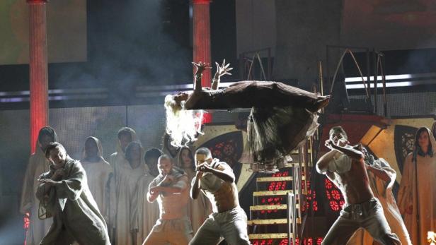 Eine Frau schwebt während einer Bühnenperformance über einer Gruppe von Tänzern.