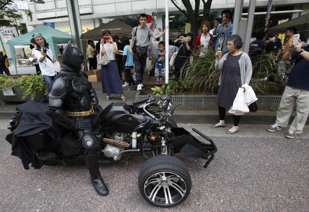 Ein als Batman verkleideter Mann sitzt auf einem Batcycle, während Passanten Fotos machen.