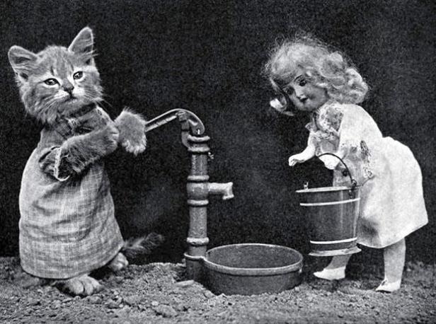 Eine Katze in Kleidung bedient eine Wasserpumpe, während eine Puppe einen Eimer hält.