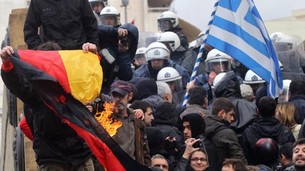 Bei einer Demonstration wird eine deutsche Flagge verbrannt, während Polizisten mit griechischen Flaggen im Hintergrund stehen.