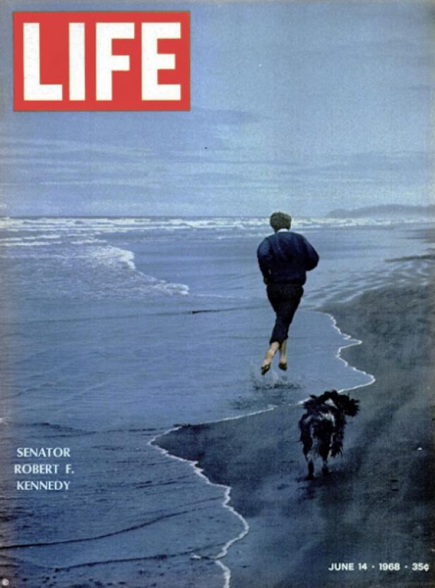 Senator Robert F. Kennedy läuft barfuß mit einem Hund am Strand entlang.