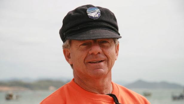 Ein Mann mit Kapitänsmütze und orangefarbenem Pullover lächelt in die Kamera.